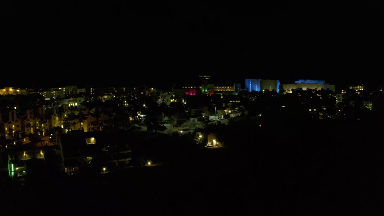 Night view of Los Cabos, Mexico, showing a low band of lit buildings, dark foreground trees, blue-lit structures in the distance, and no visible people.