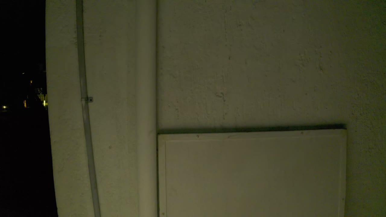 One second in, the Los Cabos frame holds on the white stucco surface, with the vertical rod still near the left edge, a dark opening on the right, and no visible people.