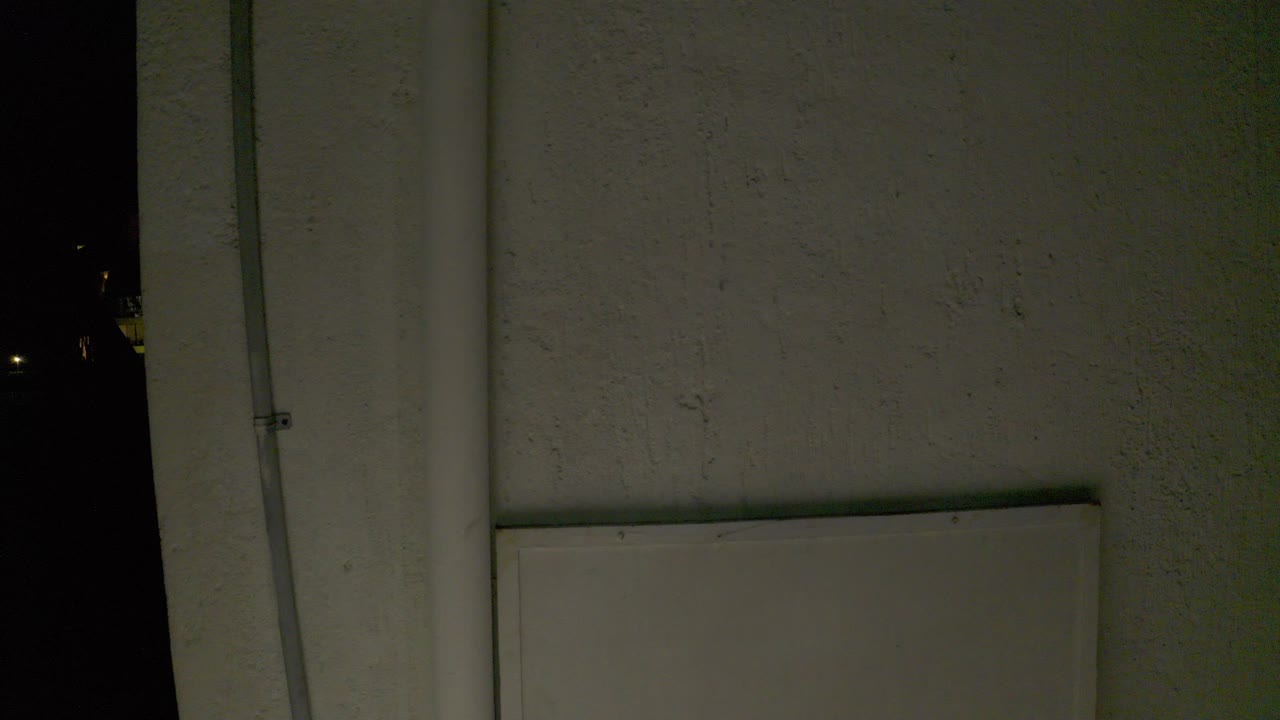 At 0:00 in Los Cabos, a dim pale wall fills the frame with a vertical conduit at left, black night space at the edges, and no visible people.