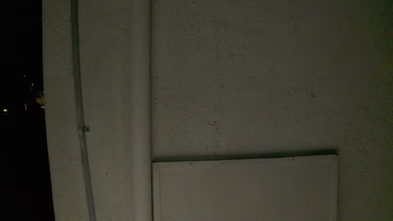 At 0:01 in Los Cabos, the pale stucco wall remains central, the left-side conduit is visible, and a few tiny lights appear in the surrounding darkness with no visible people.