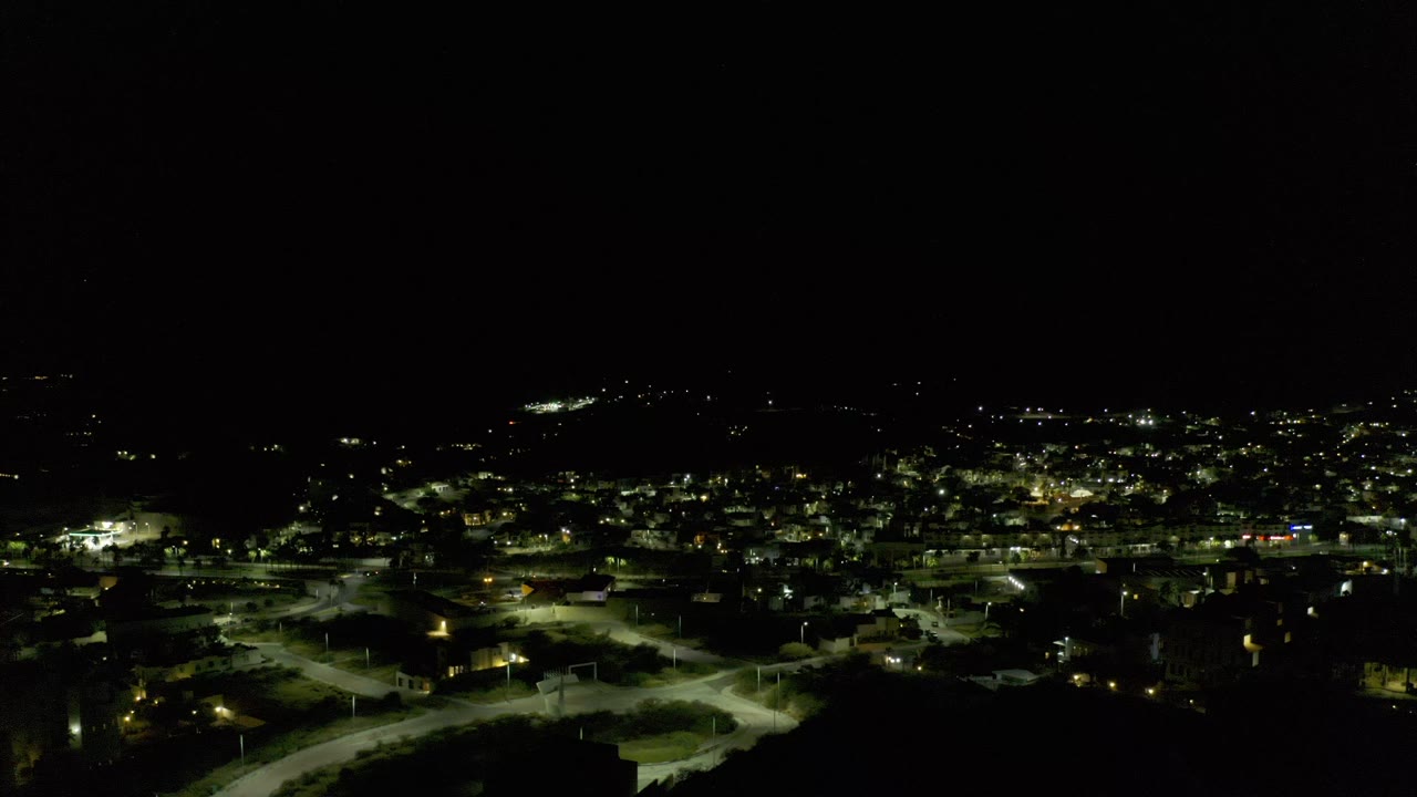 Wide nighttime aerial view over Los Cabos, Mexico, with bright street grids and scattered building lights beneath a black sky, no visible people.