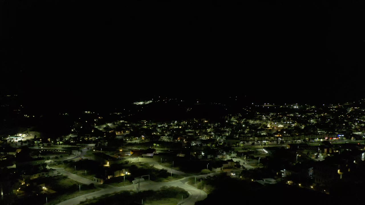 A high night view over Los Cabos shows a road grid, bright streetlamps, scattered buildings, and distant light clusters, no visible people.
