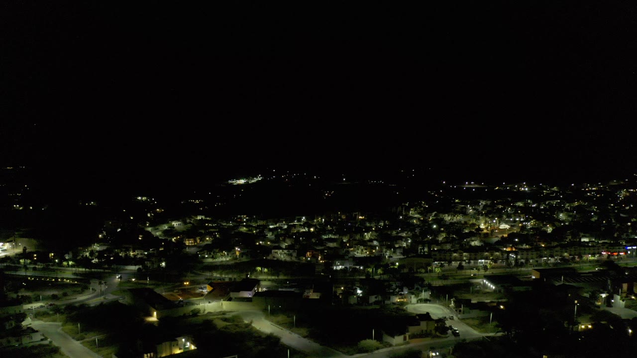 Opening nighttime aerial frame over Los Cabos, Mexico, showing illuminated neighborhood roads and scattered buildings under a large black sky, with no visible people.