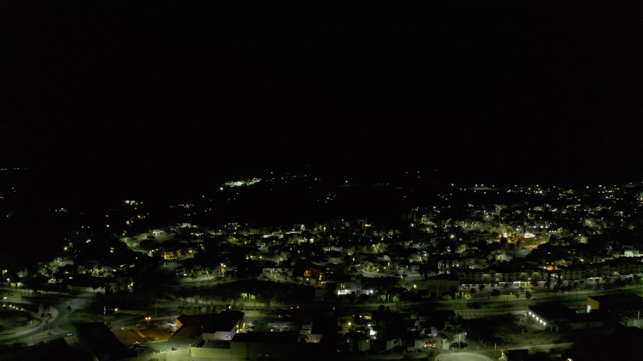 Opening frame of Los Cabos at night from an elevated aerial view, with scattered streetlights and low buildings across the lower frame and no visible people.