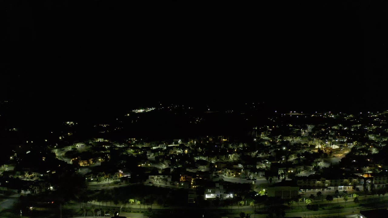 Los Cabos nighttime aerial frame showing a dense cluster of yellow-white lights, roads, and low buildings under a black upper frame, no visible people.