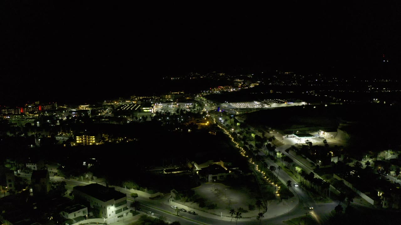 Opening frame of a Los Cabos, Mexico night aerial with a curving lit road in the foreground, clusters of buildings, distant city lights, and no visible people.