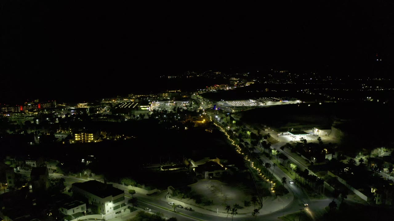 Second frame of the Los Cabos night aerial, showing the same elevated view with a brighter foreground road bend, scattered buildings, distant light clusters, and no visible people.