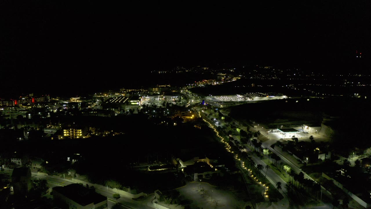 Wide night aerial over Los Cabos, Mexico with dark sky, illuminated streets, low buildings, traffic lights, and no visible people.