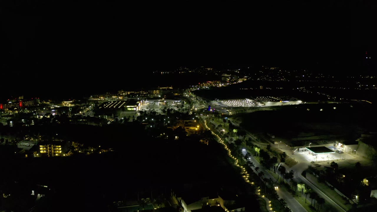 Los Cabos night scene from an elevated angle, showing a bright rectangular field or court in the lower right, lit roads through the center, and no visible people.