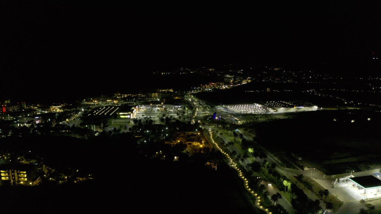 Opening aerial frame over Los Cabos at night with illuminated roads, distant city lights, dark foreground land, and no visible people.