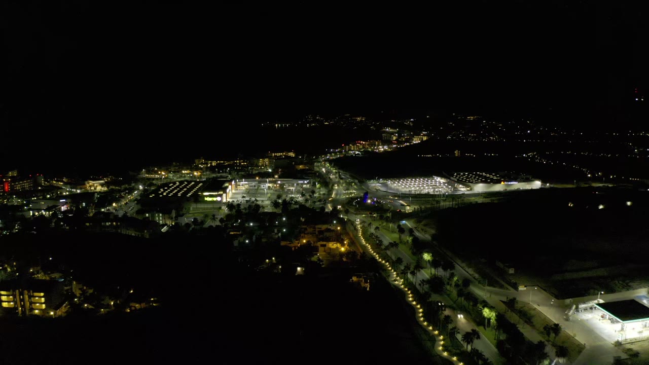 Los Cabos nighttime aerial view with a bright roadway running through the frame, lit parking areas to the left, a large illuminated lot to the right, and no visible people.