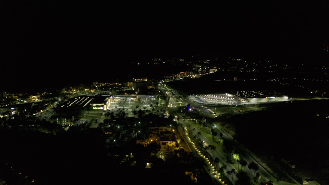 Opening night aerial over Los Cabos, Mexico, with bright roads, illuminated buildings, dark foreground rooftops, and no visible people.