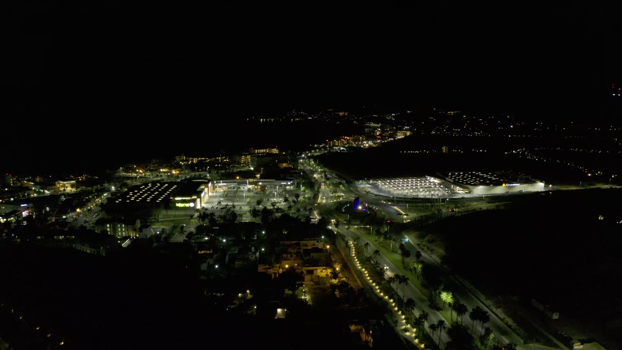 Night overhead view in Los Cabos, Mexico, showing a bright central building area, rows of parking-lot lights, dark surrounding streets, and no visible people.