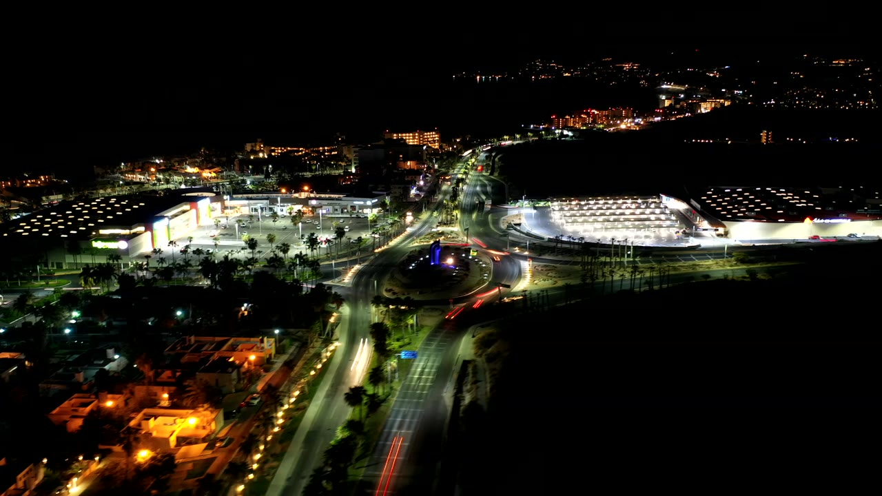 Opening frame of Los Cabos at night with bright roadways, a large illuminated complex, distant hillside lights, and no visible people.