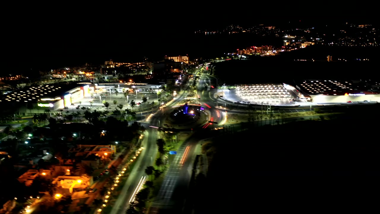 Los Cabos night frame showing bright building facades, road light trails, parking lots, palm-lined streets, and no visible people.
