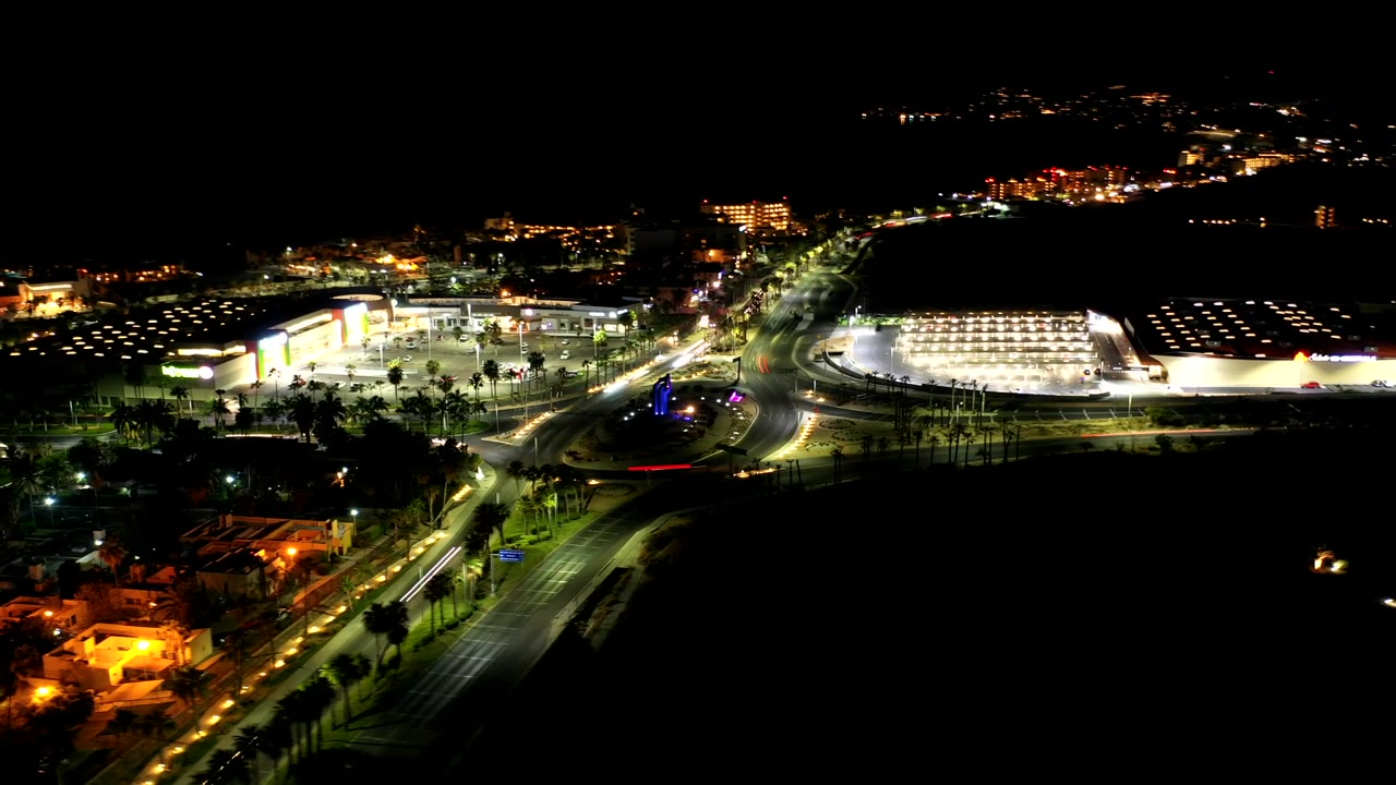 Wide night aerial over roads, parking areas, and a bright building complex.