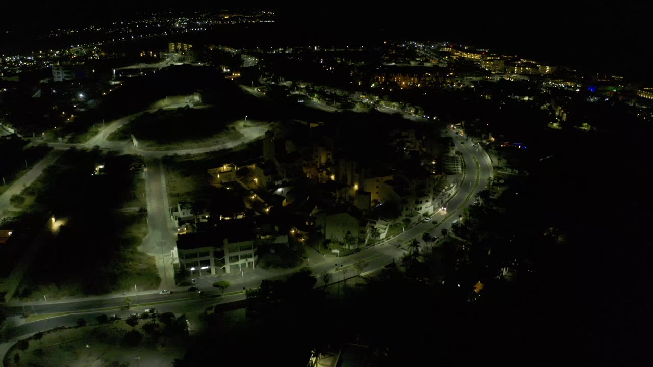 High nighttime view over Los Cabos, Mexico, showing illuminated streets, clustered buildings, dark shoreline area, and no visible people.