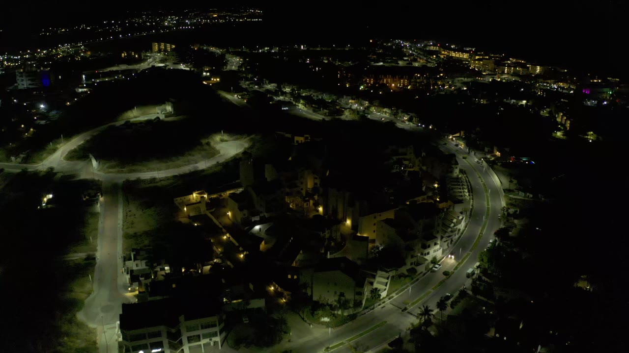 Overhead nighttime frame in Los Cabos, Mexico, showing the road curve more centrally, clusters of homes, sandy dark terrain, distant lights, and no visible people.