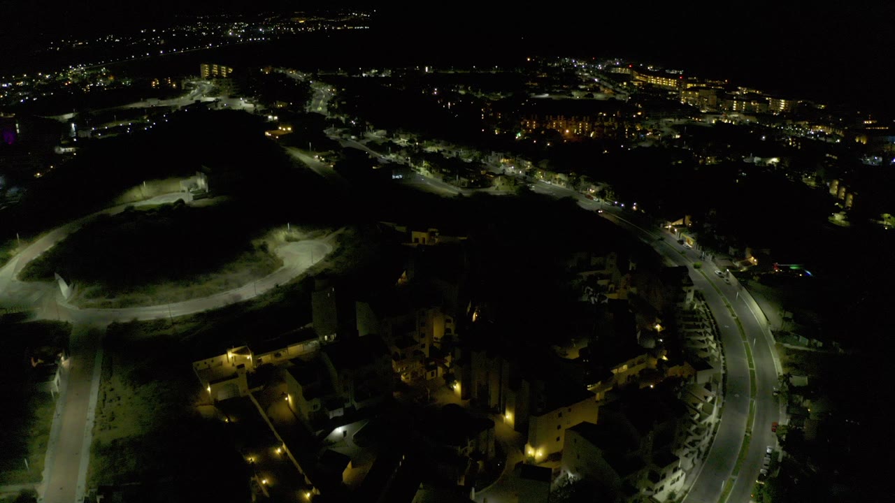 Night aerial view of a curved lit road, dark hillside, buildings, and distant city lights.
