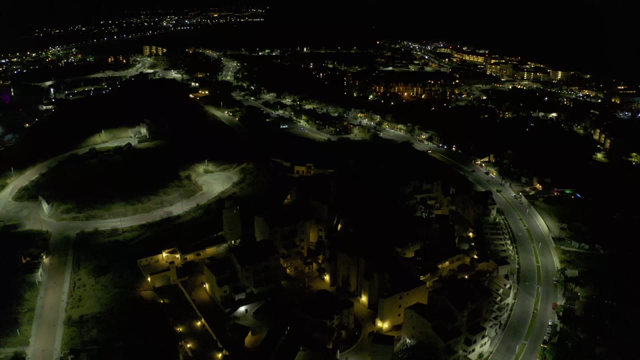High night view over Los Cabos, Mexico, showing roadway lights along the lower frame, pale paths on dark terrain, small buildings, distant city glow, and no visible people.