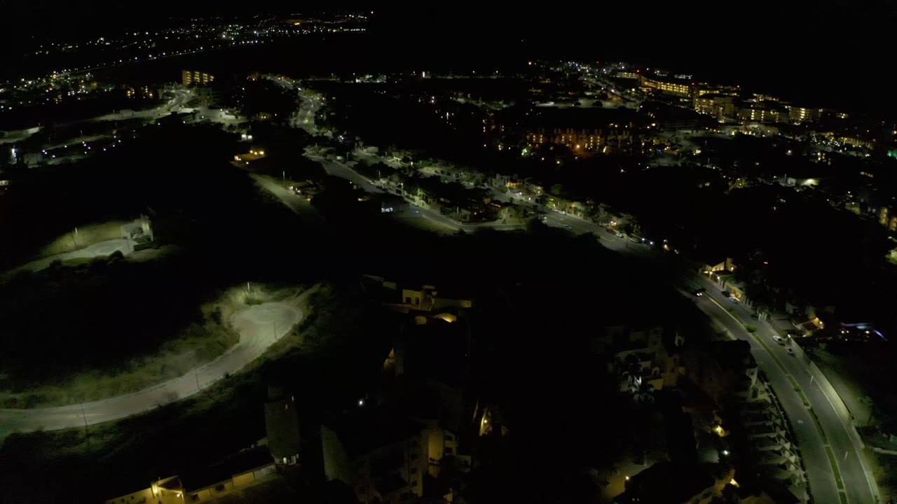Aerial frame over Los Cabos at night, showing a brighter road section, low buildings, distant city lights, and no visible people.