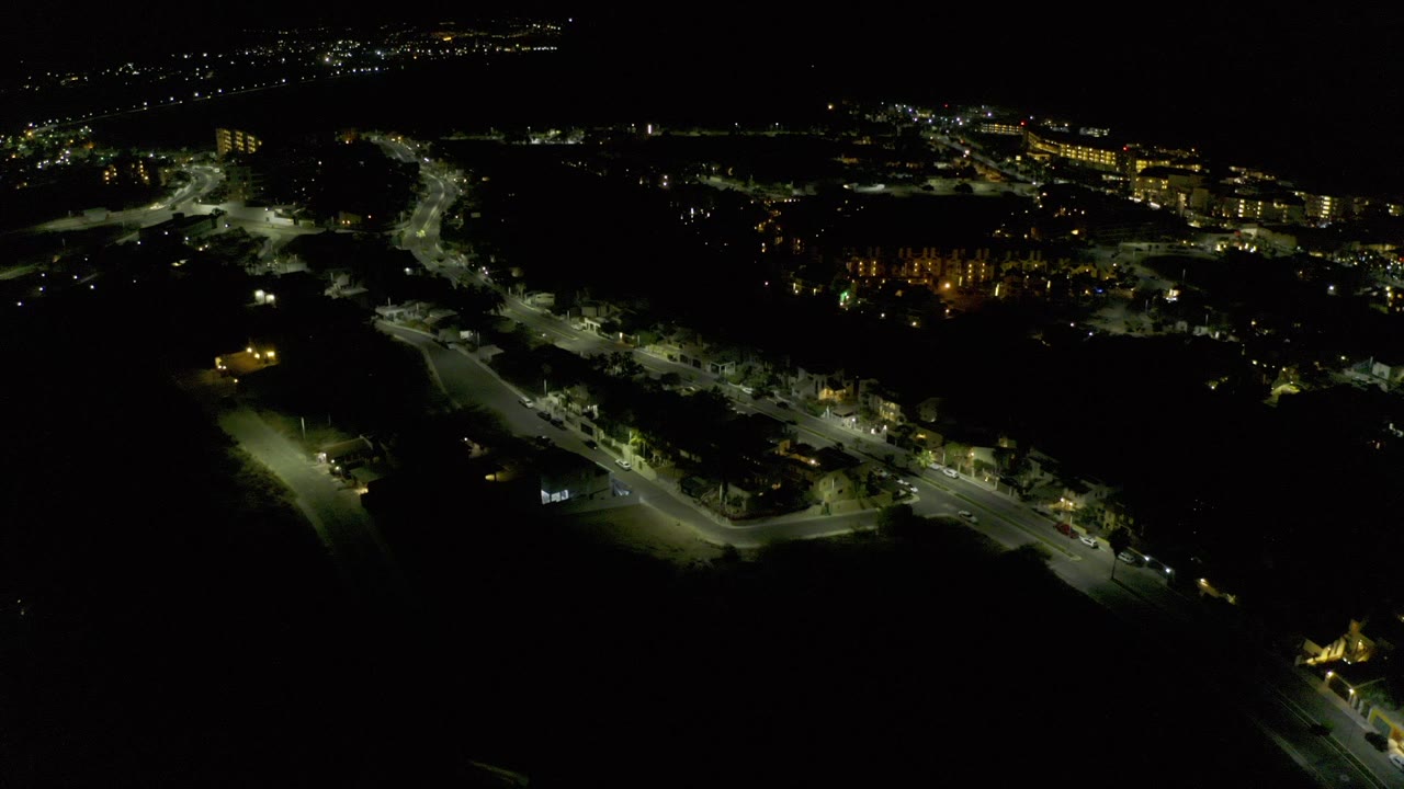 Night aerial view over Los Cabos, Mexico, with curved streets, small buildings, scattered trees, distant lights, and no visible people.
