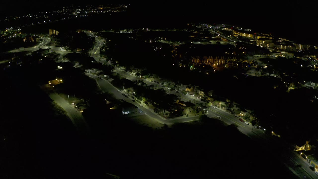 High night view in Los Cabos, Mexico, showing a brighter road junction, clusters of low buildings, dark trees, distant city lights, and no visible people.