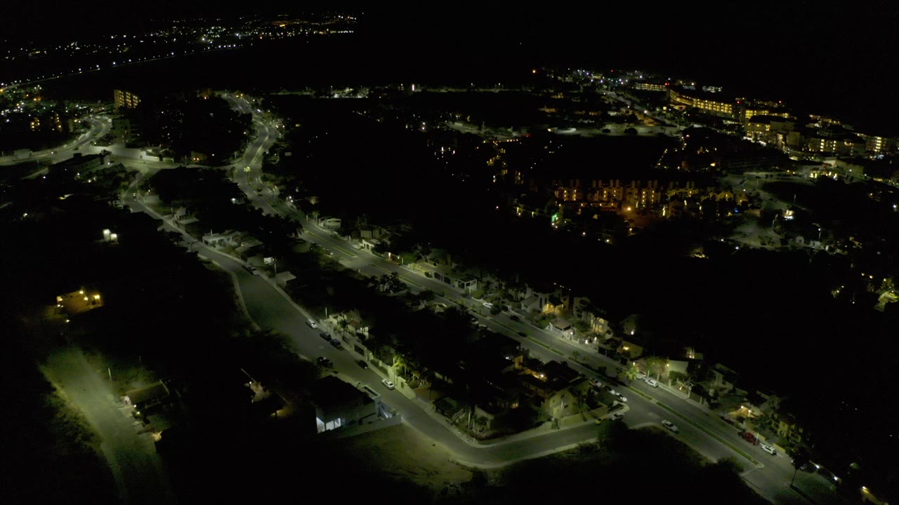 Opening night aerial over Los Cabos, Mexico, with a curving road, scattered streetlights, distant lit buildings, and no visible people.
