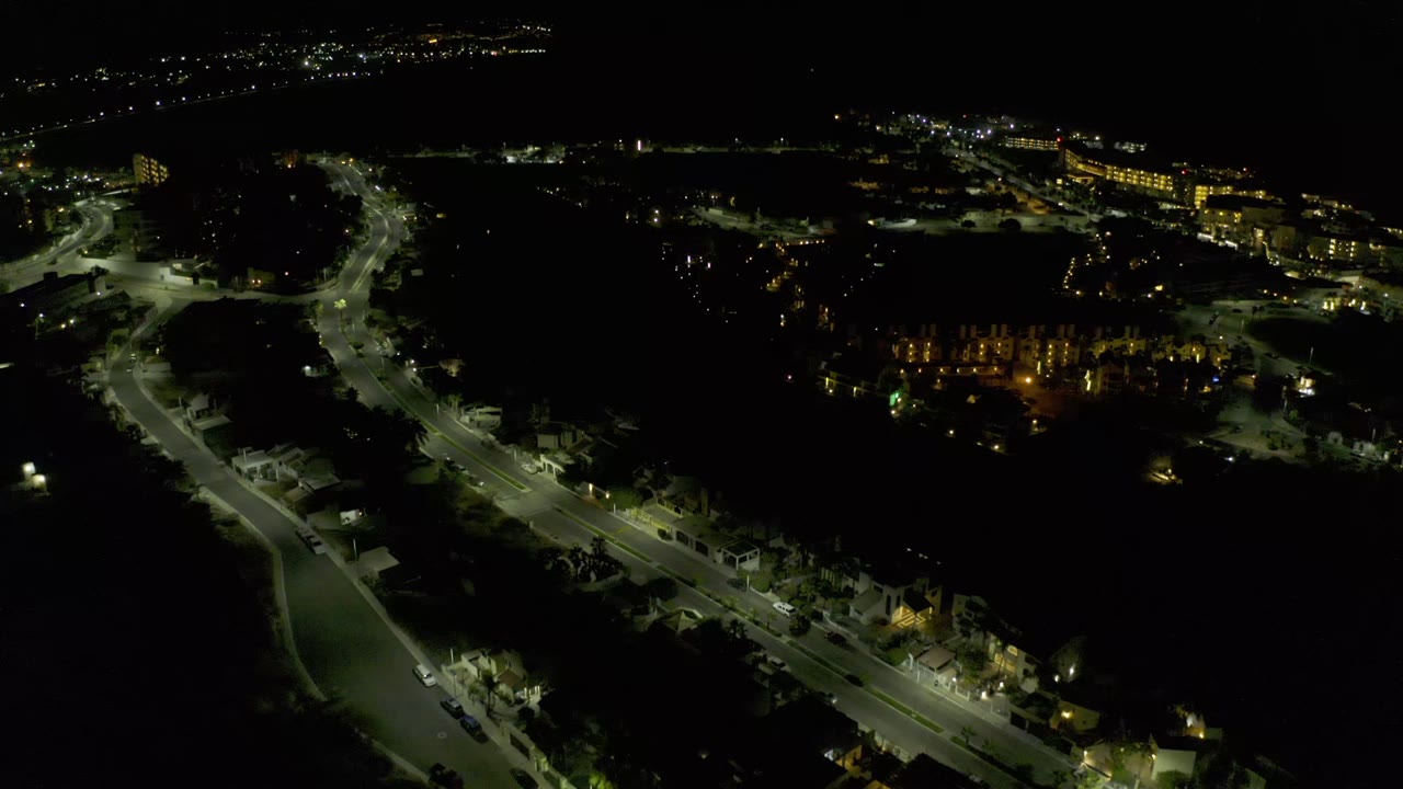 Aerial night view in Los Cabos with a bright road corridor in the foreground, clustered building lights to the right, and no visible people.