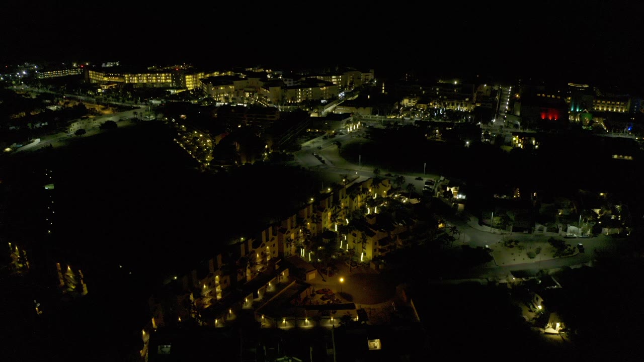 Opening nighttime aerial frame over Los Cabos, Mexico, showing warm lit buildings, parking areas, dark foreground space, and no visible people.