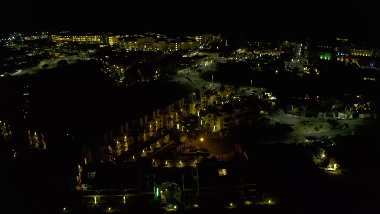 Night aerial frame in Los Cabos, Mexico, showing clustered warm lights below, open paved areas to the right, distant lights, and no visible people.