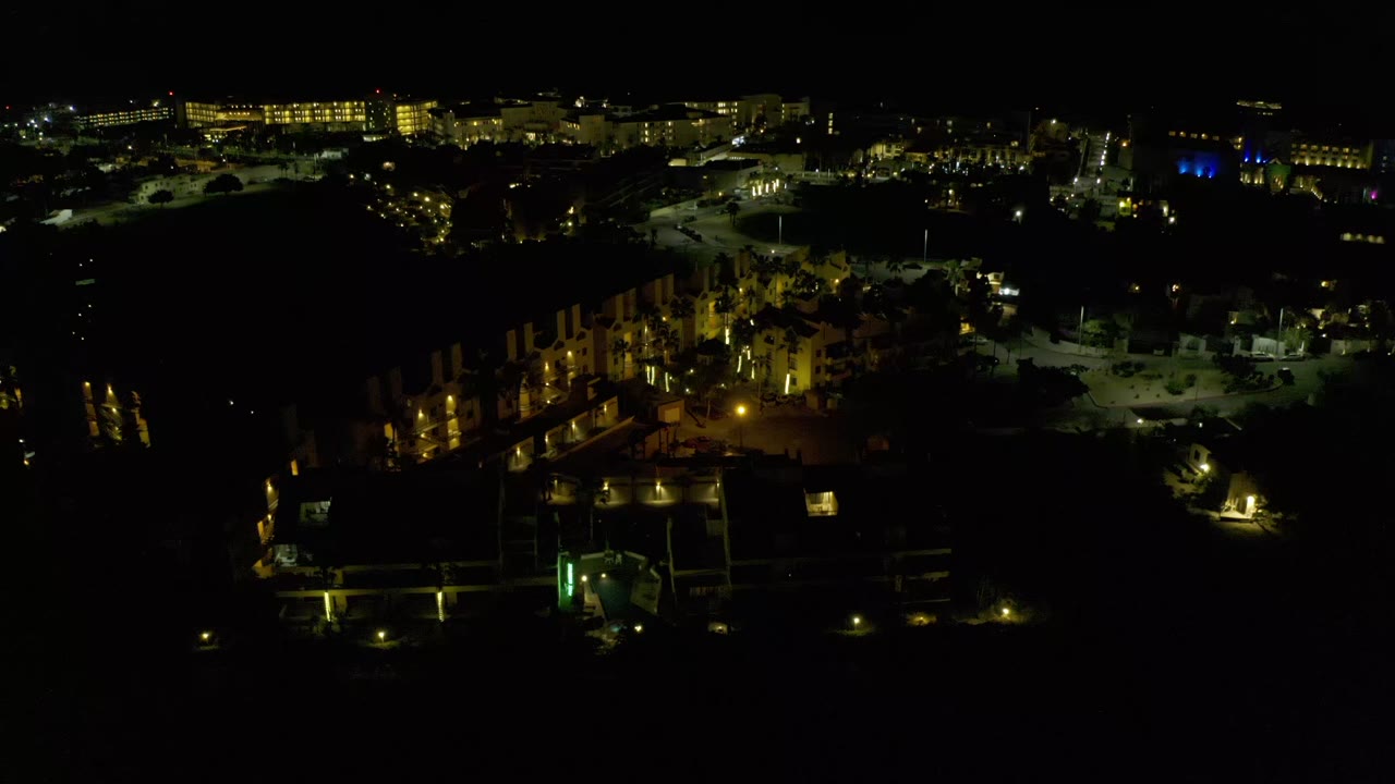 A low-light aerial view in Los Cabos, Mexico, with warm lights outlining buildings and a mostly black foreground, with no visible people.