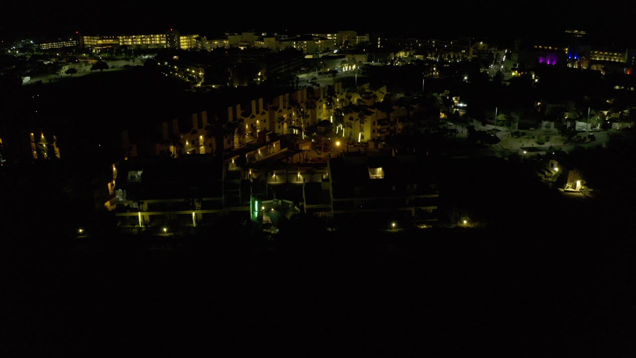 Night aerial over lit buildings and dark foreground roofs.
