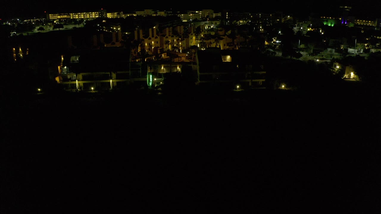 Opening frame of Los Cabos at night with a band of lit buildings across the top, scattered green and yellow lights, deep black foreground, and no visible people.