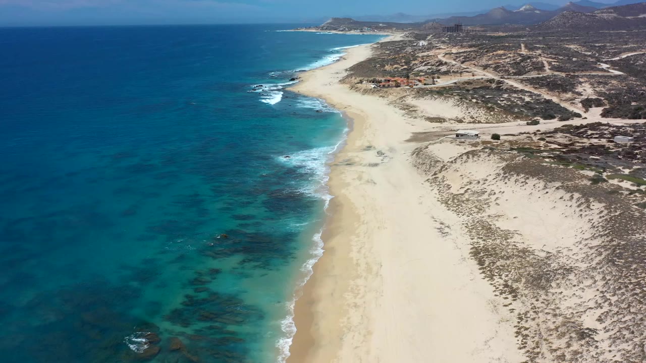 High aerial view over the Los Cabos coastline with turquoise water, pale sand, scrubby dunes, distant hills, and no visible people.