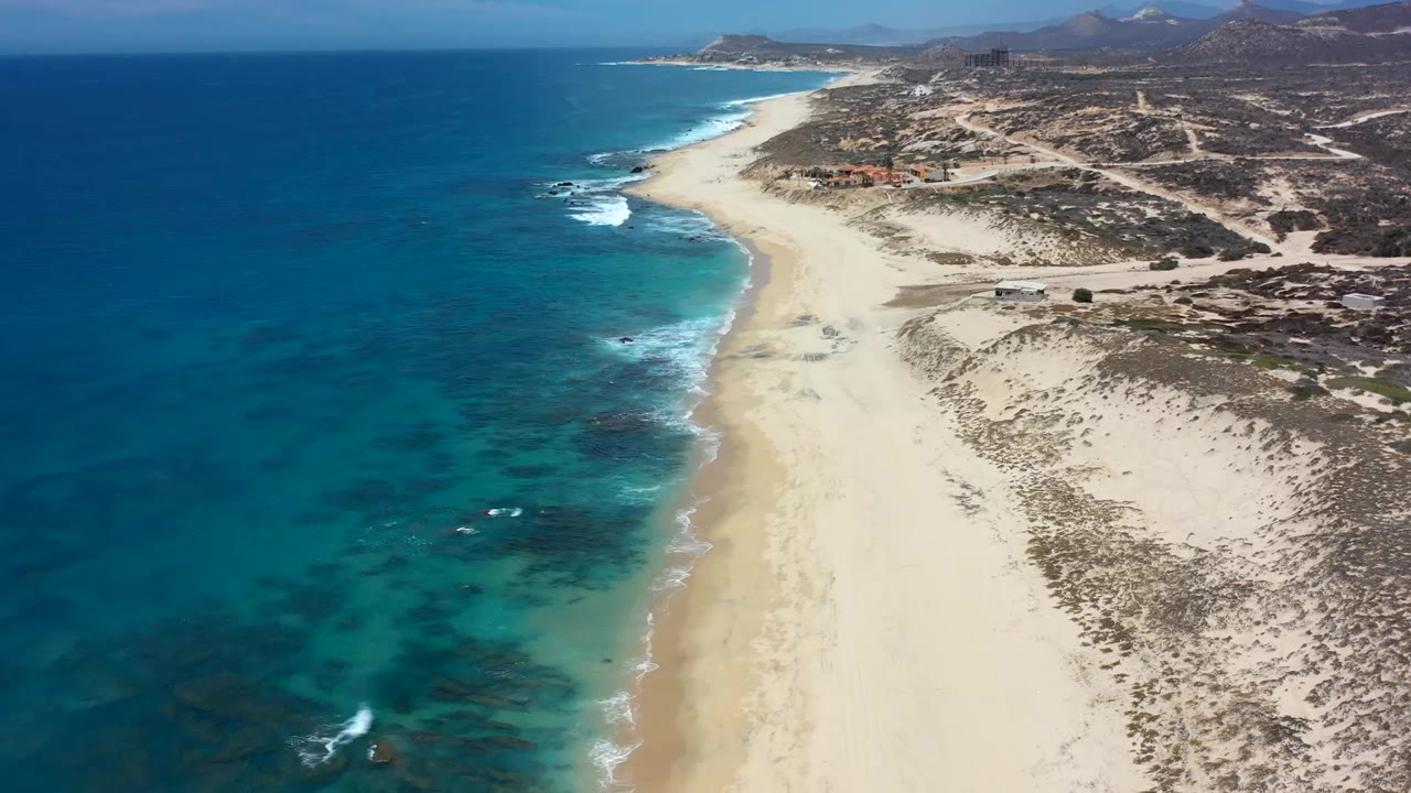Aerial frame of Los Cabos shoreline with darker reef patches under clear water, a broad empty beach, low scrubland, and no visible people.