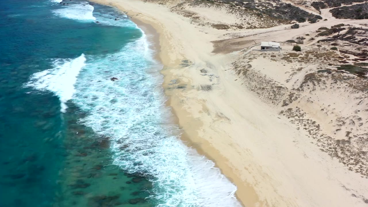 Aerial Los Cabos coastline frame with foamy waves spreading across the left side, wide sand to the right, dune scrub above, and no visible people.