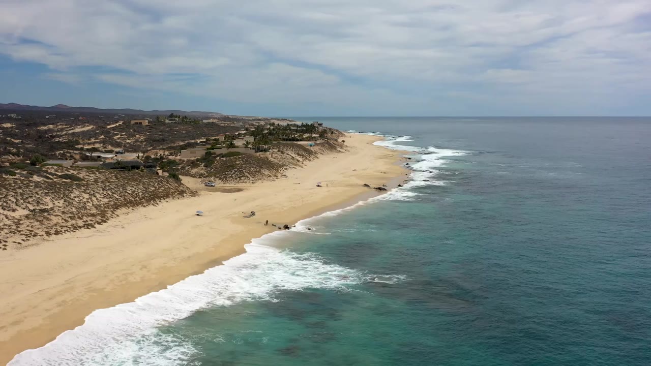 Drone view along the Los Cabos shoreline as white waves reach the sand, with desert dunes and scattered coastal structures beyond and no visible people.
