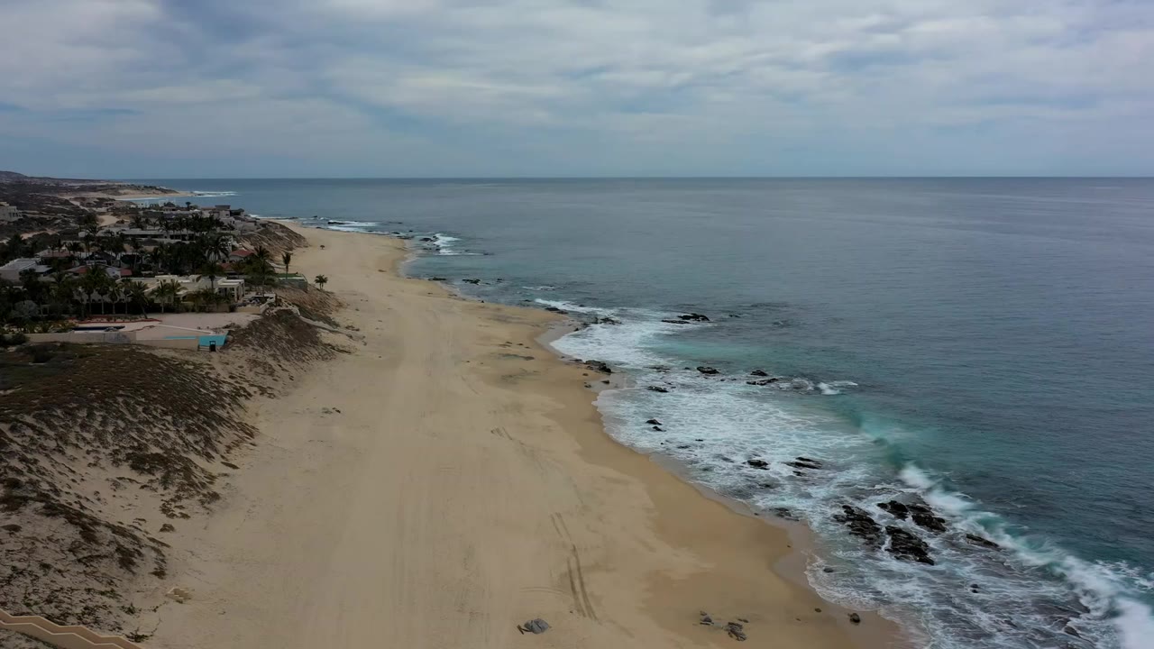 Aerial view over a Los Cabos sandy beach with white surf on dark rocks, oceanfront homes at left, an overcast horizon, and no visible people.