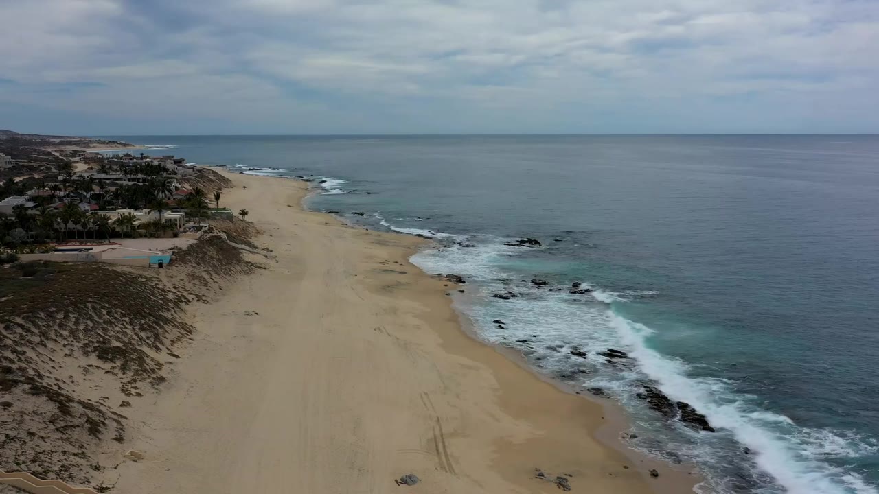 Los Cabos beach from above with a broad sand strip, gray-blue ocean, foamy waves along the rocky edge, distant shoreline, and no visible people.