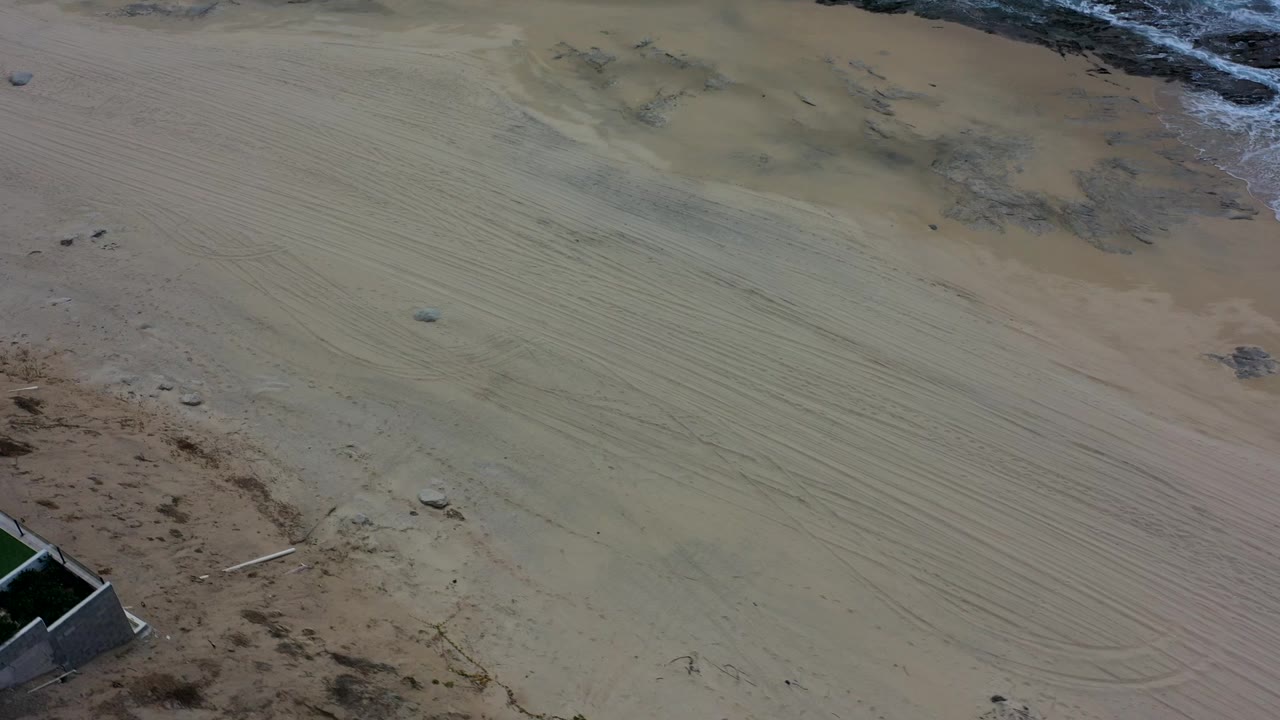 Aerial view of pale sand, dark shore rocks, and a partial beachfront wall.