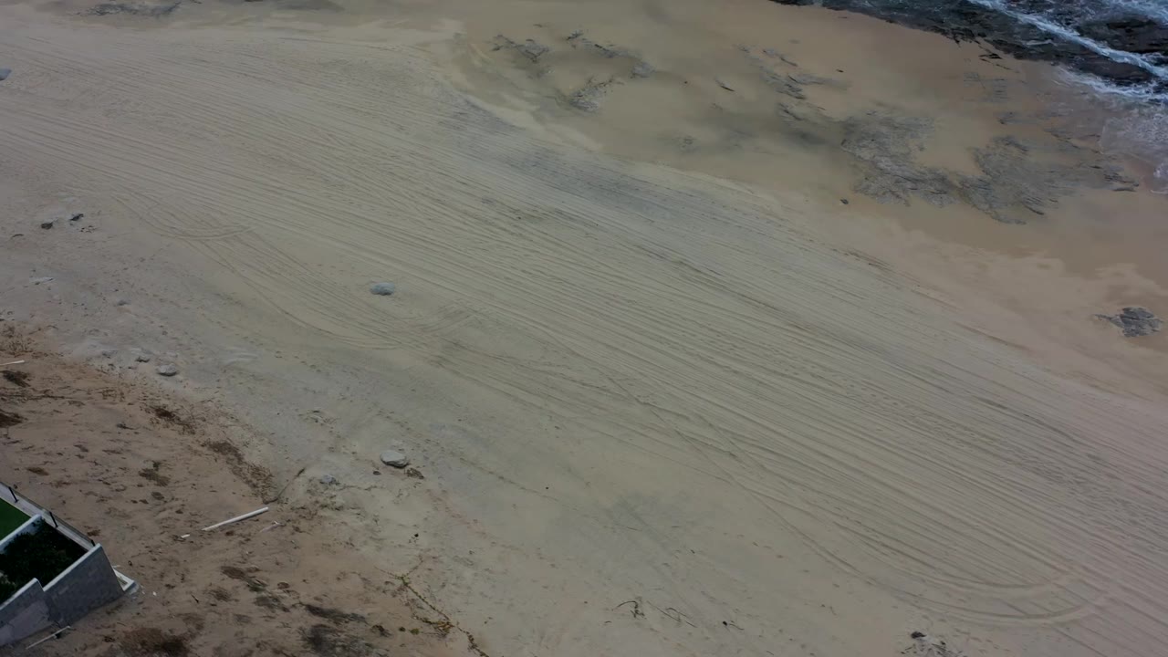 Los Cabos aerial view showing broad empty sand, scattered dark rocky patches near the waterline, visible track patterns, a property corner at lower right, and no visible people.
