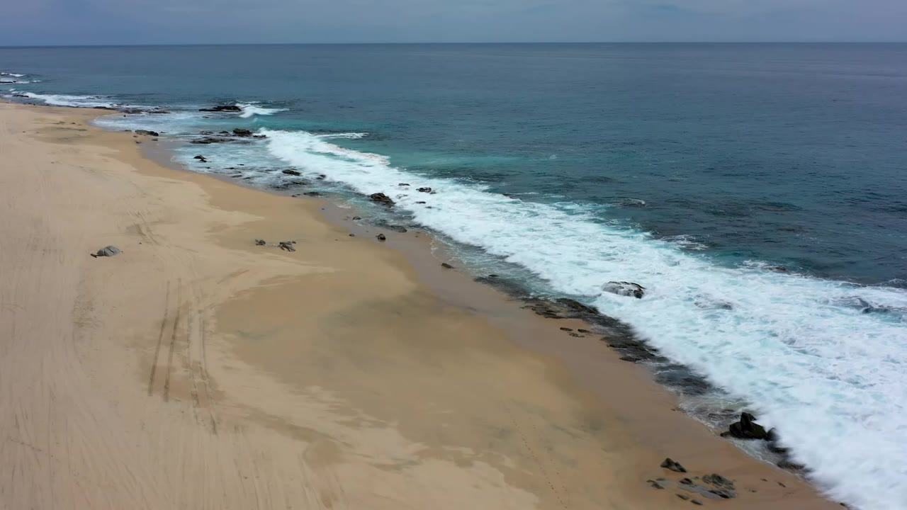 Aerial view of a Los Cabos beach where white surf curves along tan sand with dark rocks near the waterline and no visible people.