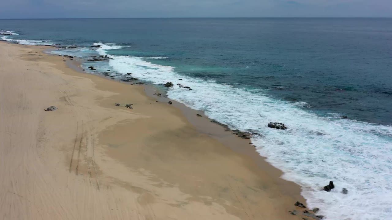 High aerial frame over Los Cabos shoreline with foamy surf on the right, open sand on the left, scattered rocks, tire tracks, and no visible people.