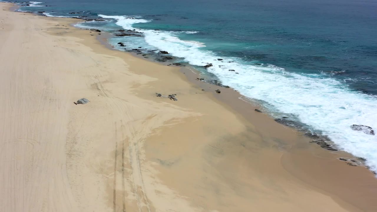 Wide aerial beach view with turquoise water and white surf rolling onto sand.