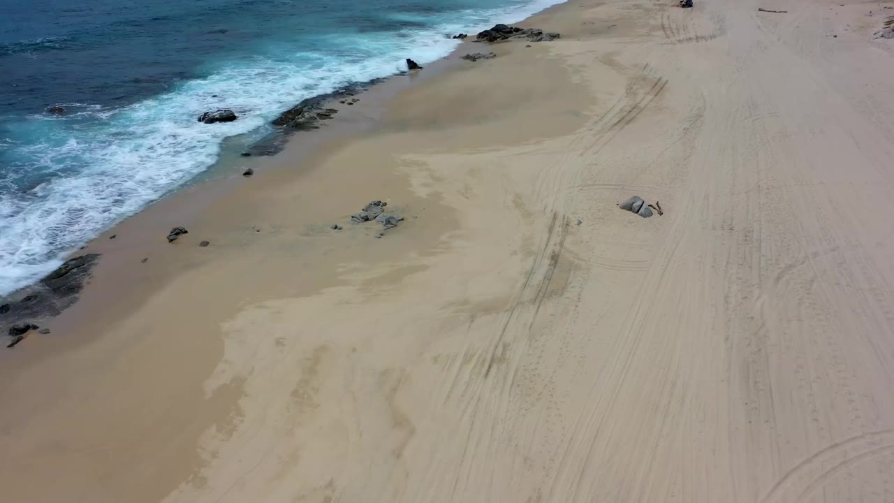 Aerial Los Cabos shoreline frame with white foam along the left edge, broad dry sand to the right, scattered rocks, faint tire marks, and no visible people.