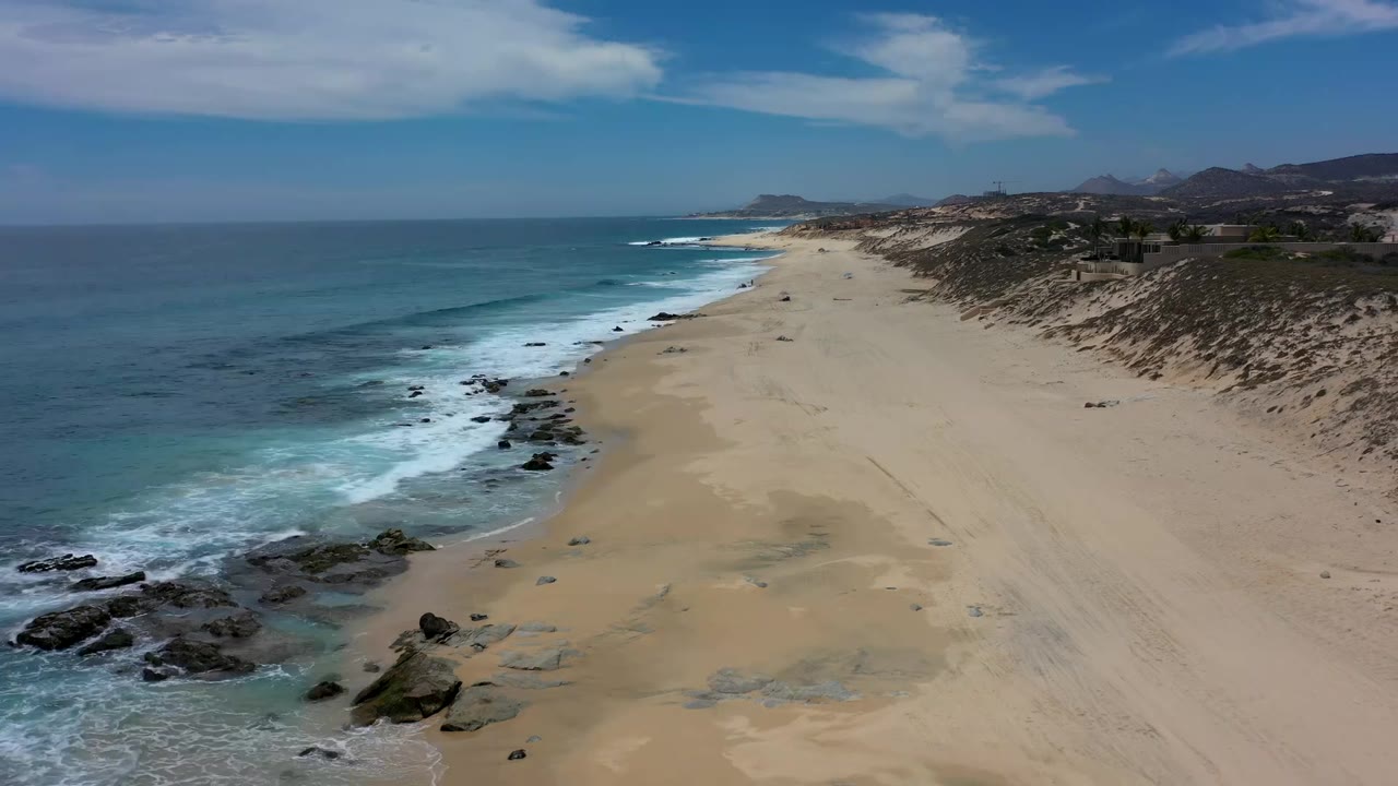 Wide Los Cabos aerial with foamy waves pushing around black rocks, pale sand stretching into the distance, low dunes on the right, and no visible people.