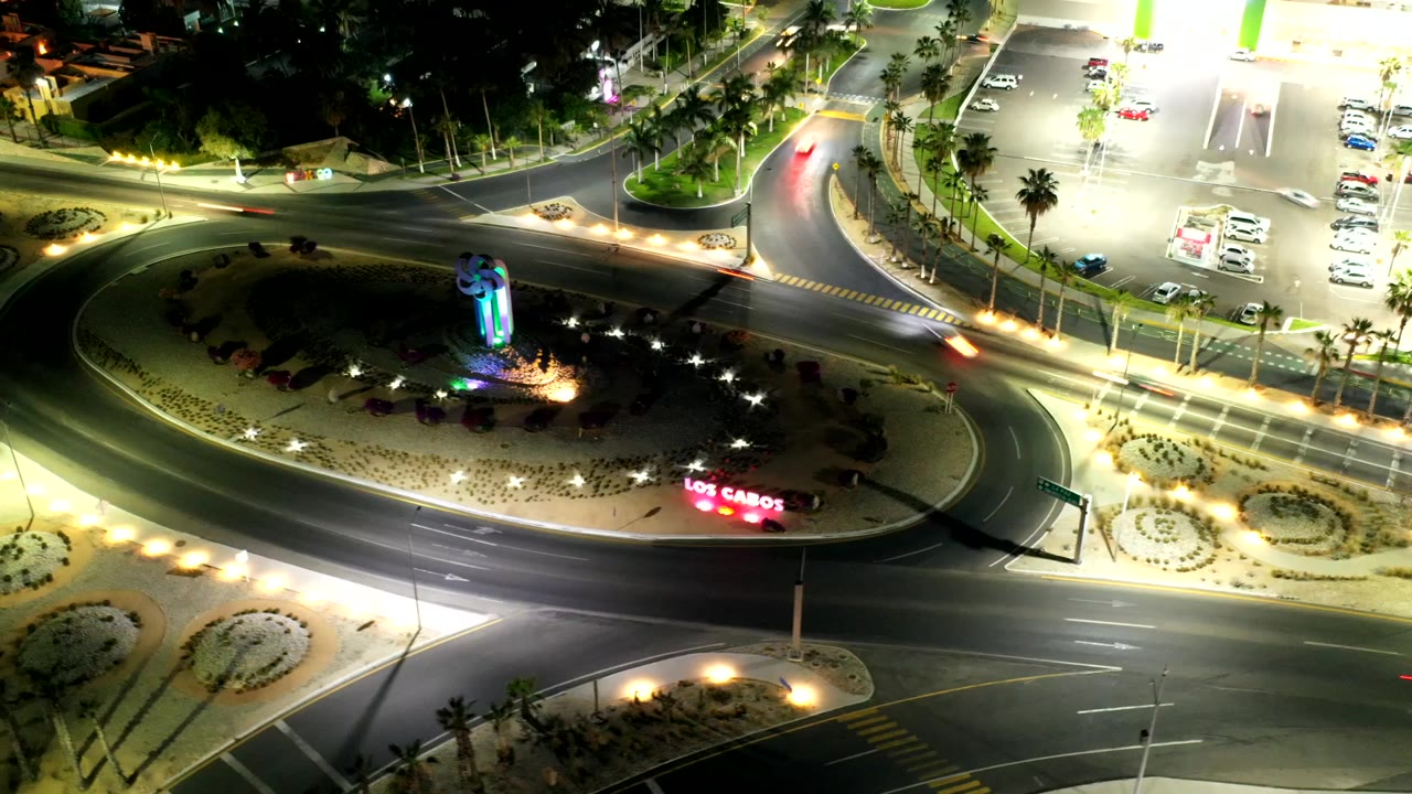 Aerial view over the Los Cabos roundabout with bright streetlights, curving traffic lanes, landscaped islands, and a parking area glowing at the upper right, with no visible people.