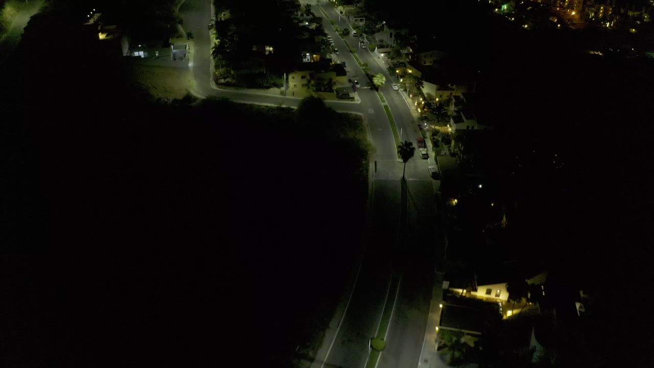 High nighttime view over Los Cabos, Mexico, showing a lit road bend, low buildings, parked cars, and deep surrounding shadows with no visible people.