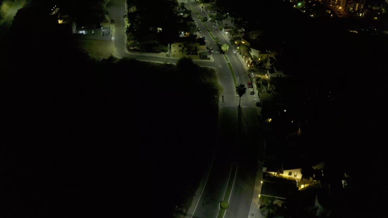 Overhead Los Cabos night frame with a curved street, scattered building lights, parked vehicles, and broad black areas around the illuminated road, with no visible people.